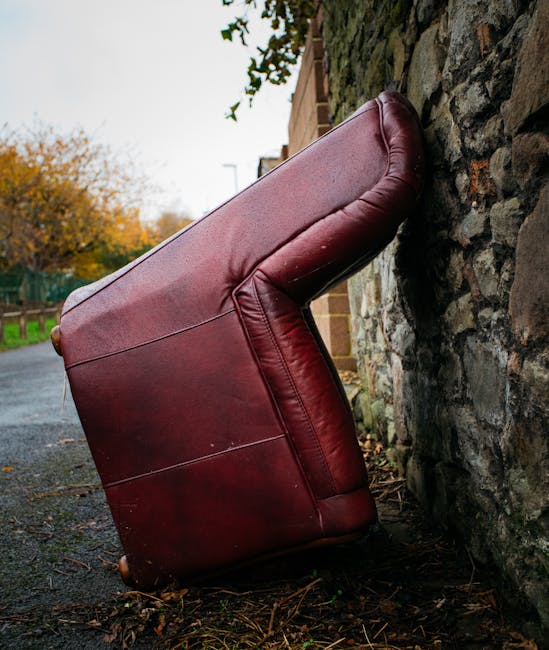 A large, burgundy leather armchair has been positioned upside down on a paved outdoor surface, leaning against a rustic stone wall. The chair's legs and underside are visible, with the upholstery showcasing a smooth, glossy finish and some creases in the material. The chair appears to be in the process of being moved or prepared for removal, and is situated near a residential building with a brick wall and a doorway, typical of home relocation or furniture transport scenarios. Behind the chair, trees with autumn foliage and a green fence can be seen under an overcast sky, indicating outdoor loading or packing activities associated with house removals. The setting suggests a typical street or driveway where furniture is loaded into a van for storage or transfer, supported by professional removal services such as those offered by Man with Van Cudham.