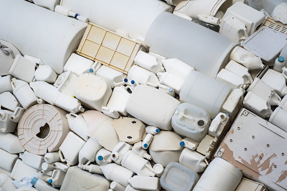 A large pile of white plastic containers, jerry cans, and bottles of various shapes and sizes, some with blue caps, stacked on top of each other indoors. Among the containers are pieces of discarded plastic parts, including a square grated cover and a round open container. The environment appears to be a waste disposal or recycling area with bright lighting. This image shows a collection of bulky plastic waste materials, relevant to home relocation and moving services that deal with waste removal, such as Man with Van Cudham, during a large item collection or clearance process involved in furniture transport and packing for relocation.