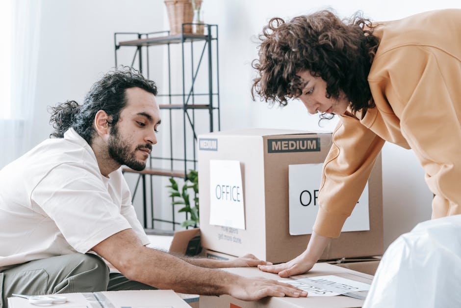Two individuals are engaged in a home relocation or packing process within a well-lit room. The man with long dark hair tied back, dressed in a white shirt and light trousers, is seated on the floor and appears to be handling packing materials or documentation related to the move. The woman with curly hair, wearing a beige top or jacket, is leaning over a large cardboard box that is labeled with various signs including 'OFFICE' and 'MEDIUM', suggesting the packing of office items for a house or office move. The box is situated on a flat surface, and her hand is placed on top of the box, possibly sealing or organizing items inside. In the background, a shelving unit and a green indoor plant are visible, indicating a typical domestic or office environment. The scene likely captures a preparation step in the moving logistics process, with [COMPANY_NAME] providing local removals and furniture transport services, supporting this packing and moving activity.