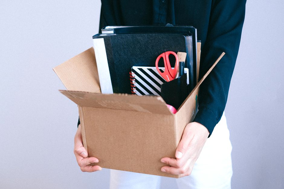 A person dressed in a dark blazer and white trousers holds an open cardboard box filled with various office supplies and personal belongings, including black binders, a spiral notebook with black and white diagonal stripes, red scissors, a wooden pencil, and a black container holding smaller stationery items. The box is positioned in front of them, and their hands support it from the bottom. The background is plain and light-colored, with natural light illuminating the scene, suggesting an indoor environment. This image illustrates the packing process involved in a home or office relocation, a service offered by companies like Man with Van Cudham, specializing in removals and furniture transport for local house and office moves, including packing and moving logistics.