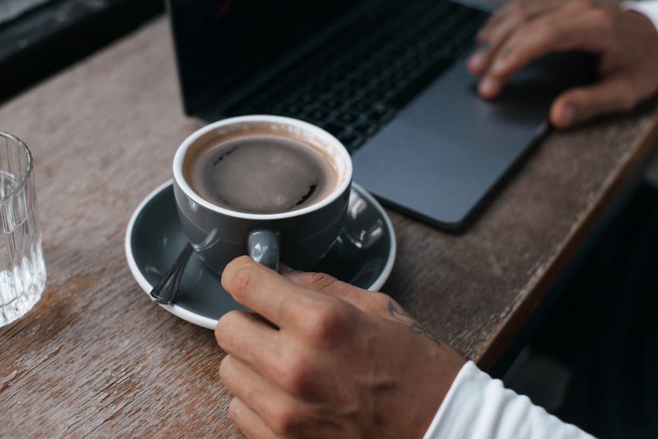 Close-up view of a white ceramic mug filled with black coffee, placed on a wooden surface with a natural grain pattern. Adjacent to the mug is an open spiral-bound notebook with lined pages, showing s