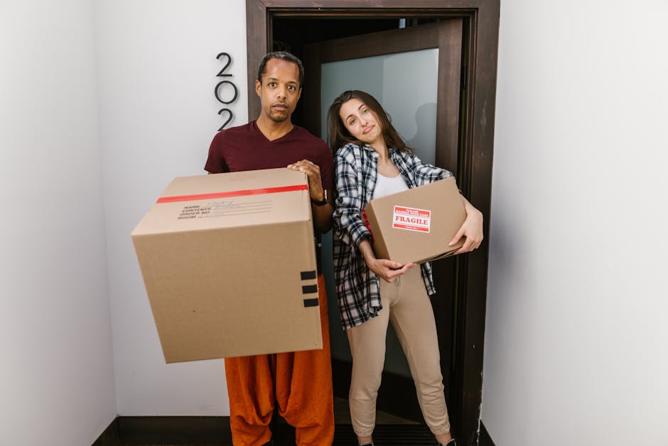 Two individuals, a man and a woman, stand inside a doorway of a residential property, holding cardboard boxes used for packing during a home relocation. The man, dressed in an orange outfit, is holding a large cardboard box with a shipping label visible on the side. The woman, wearing beige trousers and a checked shirt, holds a smaller box marked with a red and white 'FRAGILE' label. They are positioned close to each other, with the woman leaning her head towards the man, both facing the camera with neutral expressions. The background shows a plain white wall with the number '200' mounted vertically beside the door frame, which is made of dark wood. The scene appears to take place during daylight, illuminated by indoor lighting, capturing the typical packing and loading process involved in a house removal service by Man with Van Cudham, highlighting furniture transport and packing materials used during a move.
