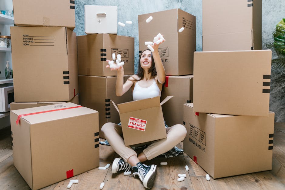 A young woman with long dark hair, sitting on a wooden floor inside a room, surrounded by numerous large cardboard moving boxes, some sealed with red tape and others open. She is inside a partially open box, smiling and tossing white packing peanuts into the air. The background shows a textured blue wall, a small cabinet with a bottle, and some green plants. The scene illustrates a home relocation process with packing and moving materials in preparation for furniture transport, reflecting the services offered by Man with Van Cudham during house removals and moving logistics.