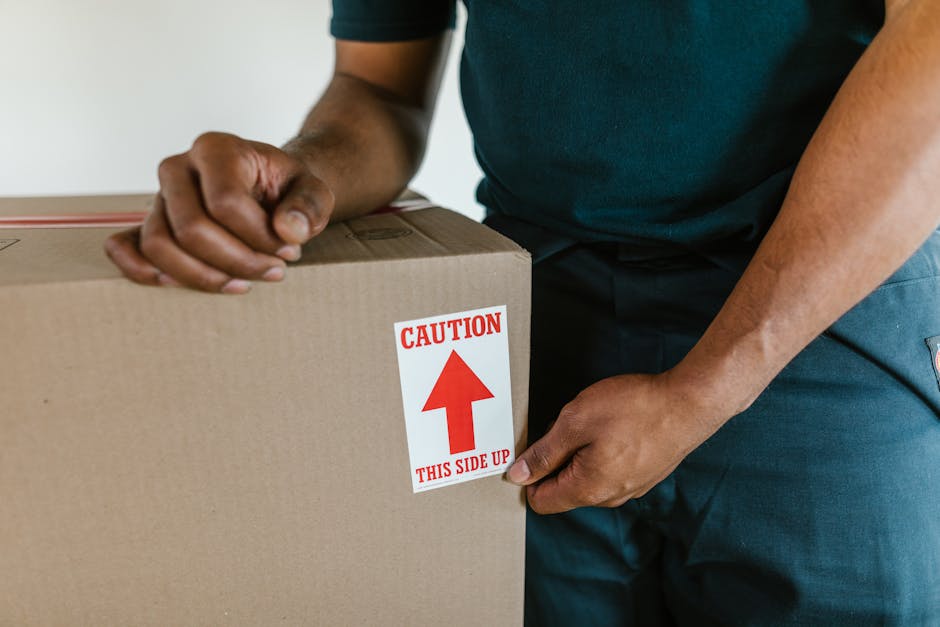 Two individuals, a man and a woman, stand inside a doorway of a residential property, holding cardboard boxes used for packing during a home relocation. The man, dressed in an orange outfit, is holding a large cardboard box with a shipping label visible on the side. The woman, wearing beige trousers and a checked shirt, holds a smaller box marked with a red and white 'FRAGILE' label. They are positioned close to each other, with the woman leaning her head towards the man, both facing the camera with neutral expressions. The background shows a plain white wall with the number '200' mounted vertically beside the door frame, which is made of dark wood. The scene appears to take place during daylight, illuminated by indoor lighting, capturing the typical packing and loading process involved in a house removal service by Man with Van Cudham, highlighting furniture transport and packing materials used during a move.