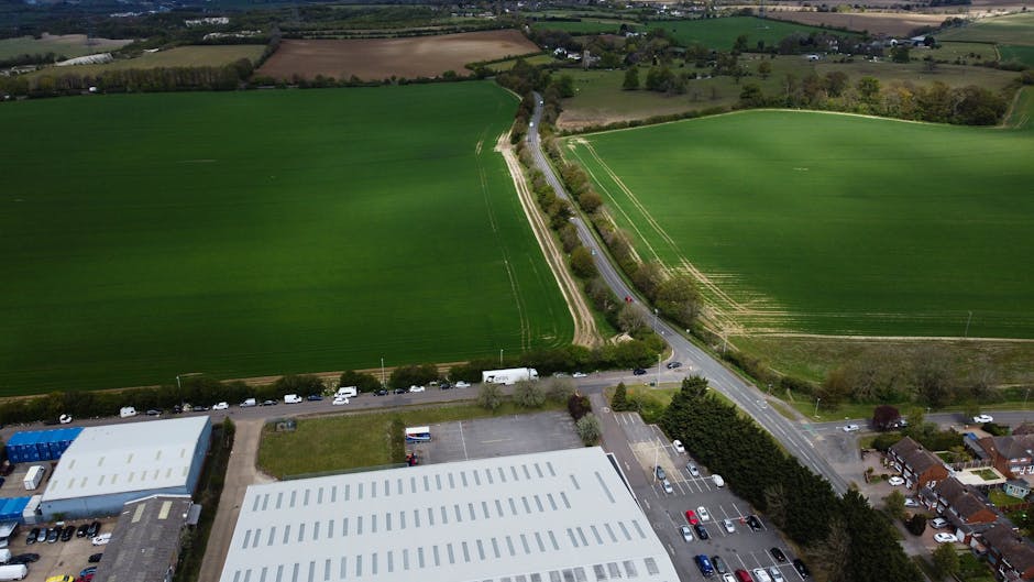 An aerial view of a rural area showing a small parking lot adjacent to a large warehouse building with a white roof. Behind the warehouse, there is a narrow road lined with trees and bushes, leading into the countryside with expansive green fields. The fields are divided by hedges and fences, with visible tractor tracks and patches of freshly ploughed land. Several cars and vans are parked along the side of the road and in the parking lot, indicating activity related to home relocation or moving services. The landscape is well-lit with natural daylight, and the scene captures the process of moving furniture and boxes in a semi-rural environment, aligning with the logistics involved in house removals and furniture transport as part of a house move, with occasional reference to packing and loading procedures.