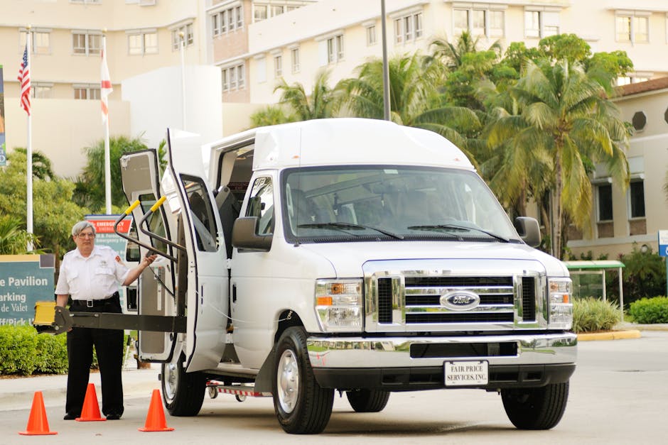 A narrow, paved alleyway between two white residential buildings with small windows and flower boxes on the right side window sills. The buildings have sloped roofs, and there are visible drainpipes along the walls. The ground is slightly wet, with a small puddle in the center reflecting light. Overhead, power lines run across, and the alley extends into the distance, where more similar buildings are visible. This setting illustrates a confined urban space suitable for detailed planning during home relocation or furniture transport, such as those handled by Man with Van Cudham, especially when navigating tight access in house removals.
