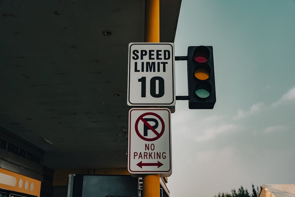 A street scene showing a traffic light with red, yellow, and green signals mounted on a yellow pole. Attached to the pole is a white speed limit sign indicating a maximum of 10 miles per hour, and a red and white no parking sign with an arrow pointing left, directing drivers to avoid parking in the area. The signs are positioned underneath a building's canopy, with the sky visible in the background featuring some clouds. This setting appears to be on a residential or commercial street, relevant to house removals and moving logistics managed by Man with Van Cudham, as part of their service on packing and loading processes for home relocation.