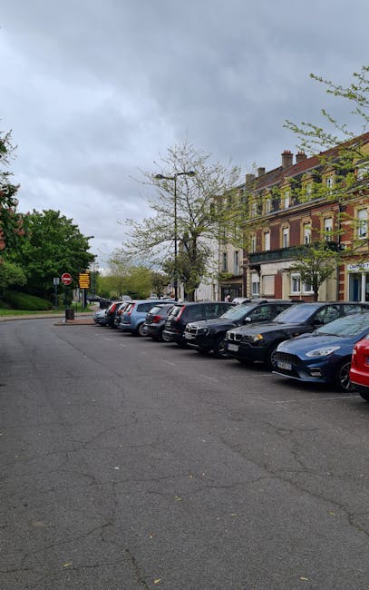 A row of parked cars, including various sedans and hatchbacks in colours such as silver, black, and blue, lined up along the curb on a residential street with a paved surface showing some cracks. Behind the vehicles, there are multi-storey Victorian-style brick buildings with large windows and decorative features, and a few trees with sparse foliage, some without leaves. The sky overhead is cloudy and overcast, creating diffuse lighting. In the background, additional street signage and greenery are visible. This scene likely depicts a parking area adjacent to a property scheduled for house removal or relocation activities. Occasionally, Man with Van Cudham offers furniture transport and home moving services, which involve careful loading and packing processes. Such tasks often include the use of trolleys, blankets, or straps, though none are visible in this image.