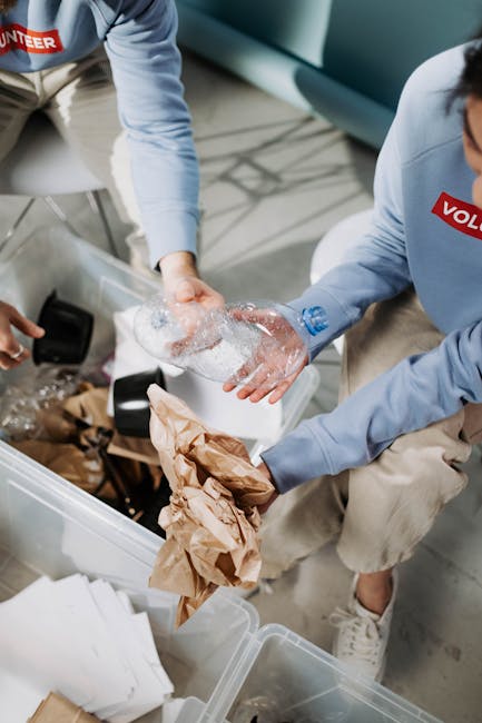 Two individuals, one wearing a light blue jacket and the other a beige sweater, are engaged in packing or preparing items for a home relocation within a modern interior space. They are seen handling a cardboard box, with one person placing or removing bubble-wrapped items inside while the other holds the box steady. Nearby, there are additional packing materials such as crumpled brown paper and white paper sheets scattered around, indicating an ongoing packing process. The scene is illuminated by soft natural or artificial light, and in the background, part of a sofa or seating area is visible, suggesting a living room or lounge environment. This activity reflects the logistical steps involved in professional removals, with [COMPANY_NAME] providing packing and transport services during a furniture and household items move. The overall setting emphasizes careful handling of belongings using packing materials and the preparation for transportation in a home relocation context.