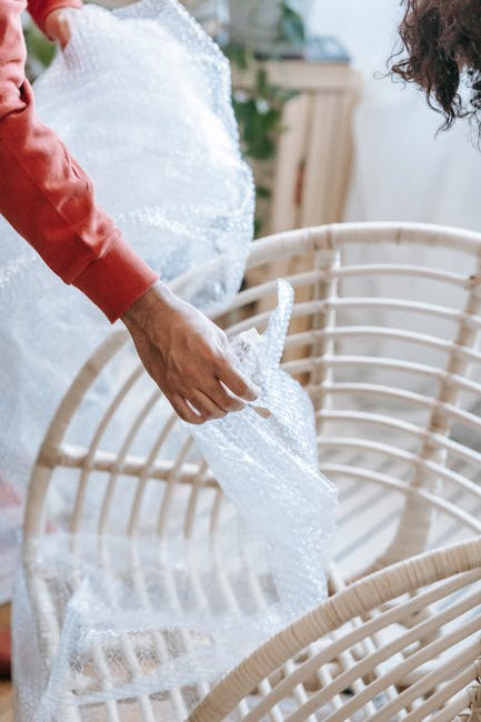 Close-up of a person wearing a red long-sleeve shirt carefully wrapping a piece of large, light-colored furniture with protective bubble wrap in an indoor setting, possibly preparing for a house move. The furniture appears to be a wooden or wicker item, situated near the doorway or inside a room with natural lighting. In the background, there are other items and a glimpse of greenery outside a window, indicating a residential environment. The person handles the protective material to ensure the furniture is securely padded for transport. This scene reflects packing and careful handling during a home relocation or furniture transport process, consistent with the services offered by Man with Van Cudham, as part of their removals and moving logistics.