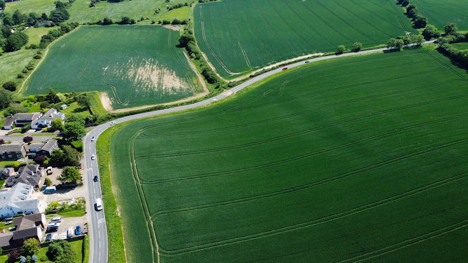 Aerial view of a rural scene showing a narrow, winding road with a few cars parked along the pavement, adjacent to a small residential area with detached houses, driveways, and gardens. Beyond the road, there are large, lush green fields separated by hedgerows, with visible crop lines and patches of exposed soil. The fields extend into the distance, demonstrating a typical countryside landscape. The image is brightly lit with natural daylight, and the overall scene captures the peaceful, open environment typical of a village setting like Cudham. This scenery supports understanding of local transport routes and rural locations relevant to house removals or moving services, as provided by companies like Man with Van Cudham, during home relocation or furniture transport in the area.