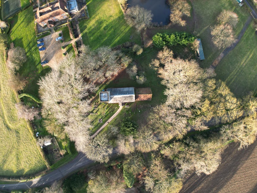 An aerial view captured during daytime showing a small, rectangular building with a flat roof and a brick extension, situated among numerous leafless and leafy trees on a large, well-maintained green lawn. A narrow, curved paved driveway leads to the building, surrounded by open grassy areas and gardens. To the top left, a gravel parking area with several parked cars is visible, adjacent to a pathway that connects to nearby residential properties. In the background, additional trees, pathways, and landscaped gardens are visible. This scene depicts a peaceful countryside setting consistent with a house relocation process, where the surrounding environment facilitates loading or unloading activities as part of a professional removals service, such as those offered by Man with Van Cudham.