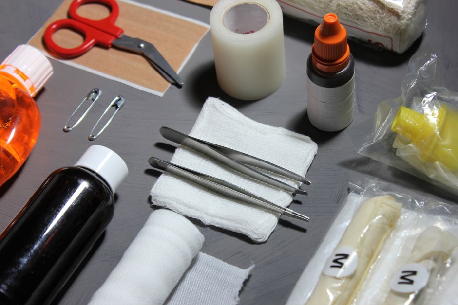 An assortment of medical and first aid supplies arranged on a grey surface, including a roll of medical tape, a pair of stainless steel forceps and scissors, a folded white towel, a small orange-capped bottle, a larger black bottle, and various plastic-wrapped items such as gauze and bandages. The items are positioned near a pair of orange-handled scissors, metal safety pins, and a few other small containers, all set up in an indoor environment likely related to packing or handling medical or hazardous materials during a house removal or relocation process. Some items are placed on a brown backing sheet, possibly for easy handling. The scene reflects careful preparation and organisation typical of packing hazardous or delicate items safely, relevant to the packing and moving services provided by Man with Van Cudham, particularly in the context of home relocation and furniture transport.