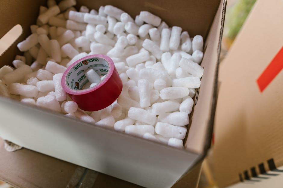 Inside a cardboard moving box filled with white packing peanuts, there is a roll of red adhesive tape with green and white branding placed on top of the packing materials. The box is positioned outdoors, near a doorway or a vehicle used for home relocation, with part of a moving van or storage area visible on the right. The scene suggests the packing and loading process involved in a house removal service, with focus on safe packaging materials for transporting fragile or hazardous items as part of furniture transport or packing and moving activities conducted by Man with Van Cudham.