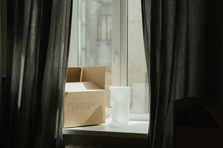 A cardboard box labeled 'CLOTHES' sits on a windowsill inside a home, surrounded by grey curtains pulled slightly apart. Next to the box, there is a white rolled-up fabric or packing material. The window is open, revealing an exterior building wall made of stone and a small part of a window from the outside. The scene is lit with natural daylight, indicating daytime, and the overall setting suggests the packing phase of a home relocation. This image is associated with furniture transport and packing during house removals, as provided by Man with Van Cudham, part of their elimination and moving services.