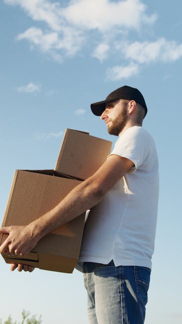 A man with a beard, wearing a black baseball cap, white t-shirt, and blue jeans, is standing outdoors under a partly cloudy sky, holding two cardboard boxes stacked one on top of the other in front of him during a home relocation process. The boxes appear to be securely sealed with packing tape. Behind him, there is a van with an open door; the scene suggests the loading or unloading phase of a house move, with the man preparing the boxes for transport. The environment is well-lit with natural daylight, and the focus is on the man, the boxes, and the vehicle, illustrating aspects of furniture transport and packing during a professional removals service, as provided by Man with Van Cudham.